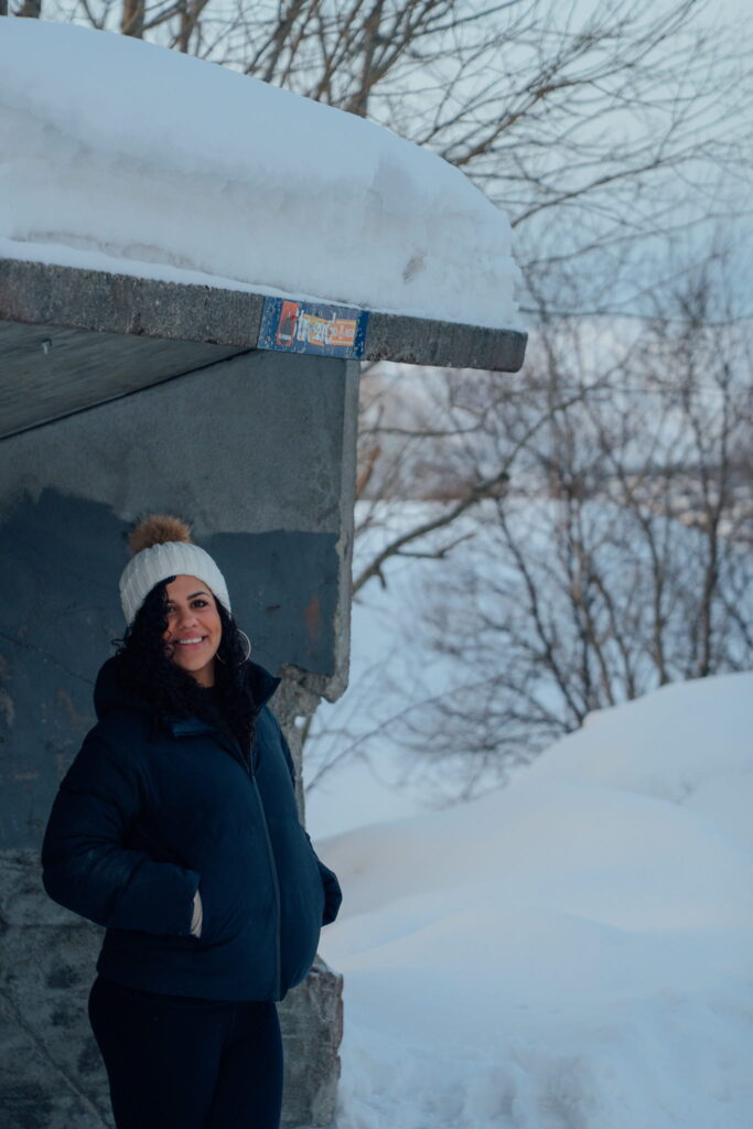 A woman in a dark winter jacket and white knit hat stands smiling under a snow-covered structure, surrounded by snow and bare trees—a perfect scene for your 3 Day Tromso Itinerary in this magical wintry landscape.