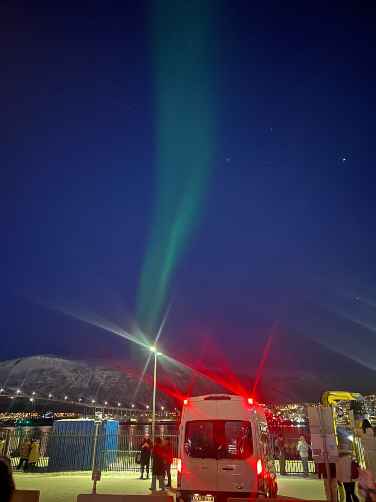 A green aurora lights up the night sky above snowy mountains, as people following a 3 Day Tromso Itinerary stand near a white van and a brightly-lit bus at a parking area with streetlights and a fence.