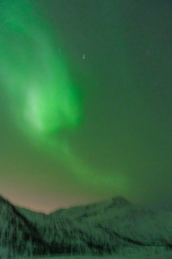 A vivid green aurora borealis lights up the night sky above snow-covered mountains, casting a soft glow over the wintry landscape—a magical highlight on any 3 Day Tromso Itinerary.