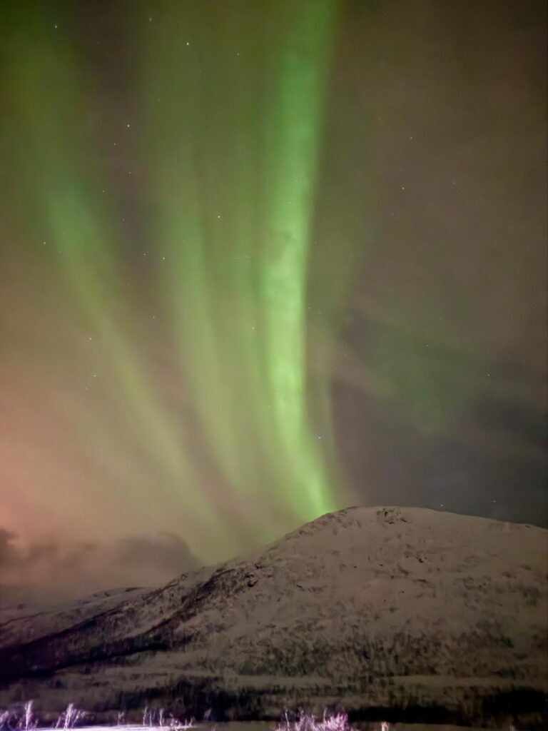 Green aurora borealis lights arc across a cloudy night sky above a snow-covered mountain landscape, creating a vibrant and ethereal scene perfect for any 3 Day Tromso Itinerary.