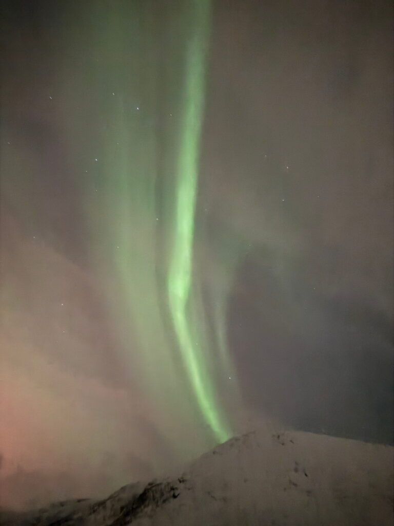 Green aurora borealis lights swirl across a cloudy night sky above a snowy mountain peak, with a few visible stars dotting the background—a breathtaking scene perfect for any 3 Day Tromso Itinerary.