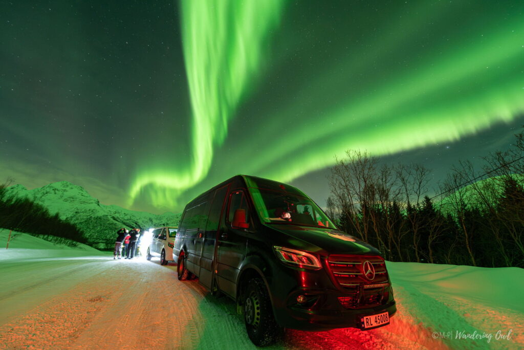 A dark van and cars parked on a snowy road under bright green Northern Lights, with mountains and trees in the background—a magical moment often included in a 3 Day Tromso Itinerary. Several people stand by, illuminated by headlights.