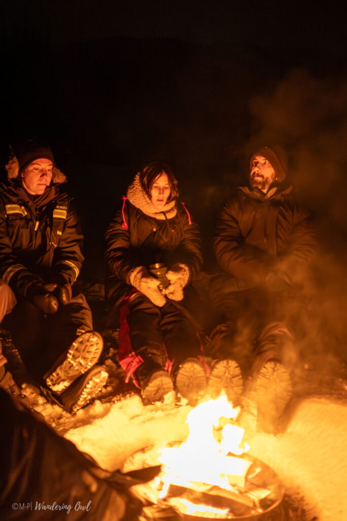 Three people dressed in winter clothing sit around a campfire at night, their faces aglow, snow on the ground—capturing the cozy spirit of a 3 Day Tromso Itinerary amid Arctic darkness.