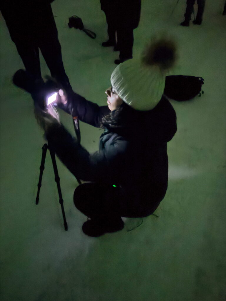 A person wearing a white knit hat with a pom-pom squats in the snow at night, adjusting a camera on a tripod. The scene captures the spirit of adventure on a 3 Day Tromso Itinerary, with others and bags visible in the dim background.