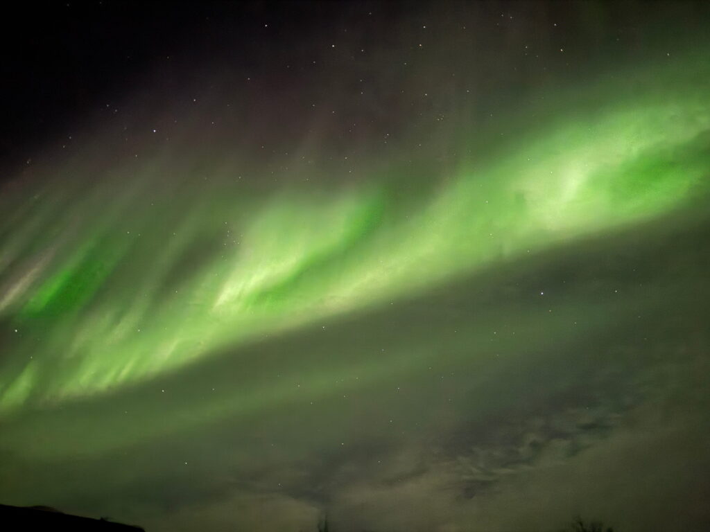 Green aurora borealis lights stretch across a dark night sky, with faint stars in the background—a magical sight to include on your 3 Day Tromso Itinerary, with wispy clouds near the horizon completing the scene.
