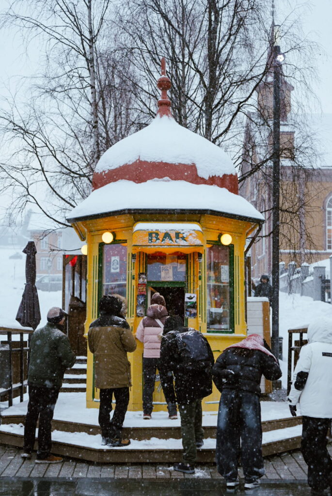 People stand in front of a small, yellow "BAR" kiosk covered in snow—an inviting stop on your 3 Day Tromso Itinerary—with snow falling and wintry trees and buildings forming the backdrop on a chilly winter day.