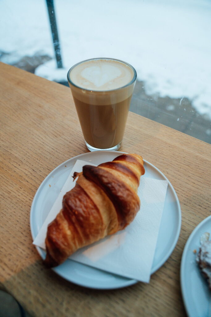 A glass of latte with heart-shaped foam sits on a wooden table next to a croissant, perfect fuel as you plan your 3 Day Tromso Itinerary, with a snowy outdoor scene visible through the window in the background.