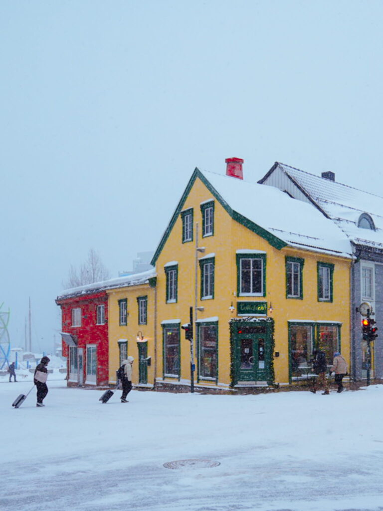 People walk with suitcases near a bright yellow and green corner building on a snowy street, as snow falls—an inviting scene for anyone planning a 3 Day Tromso Itinerary, with a red building visible in the background.
