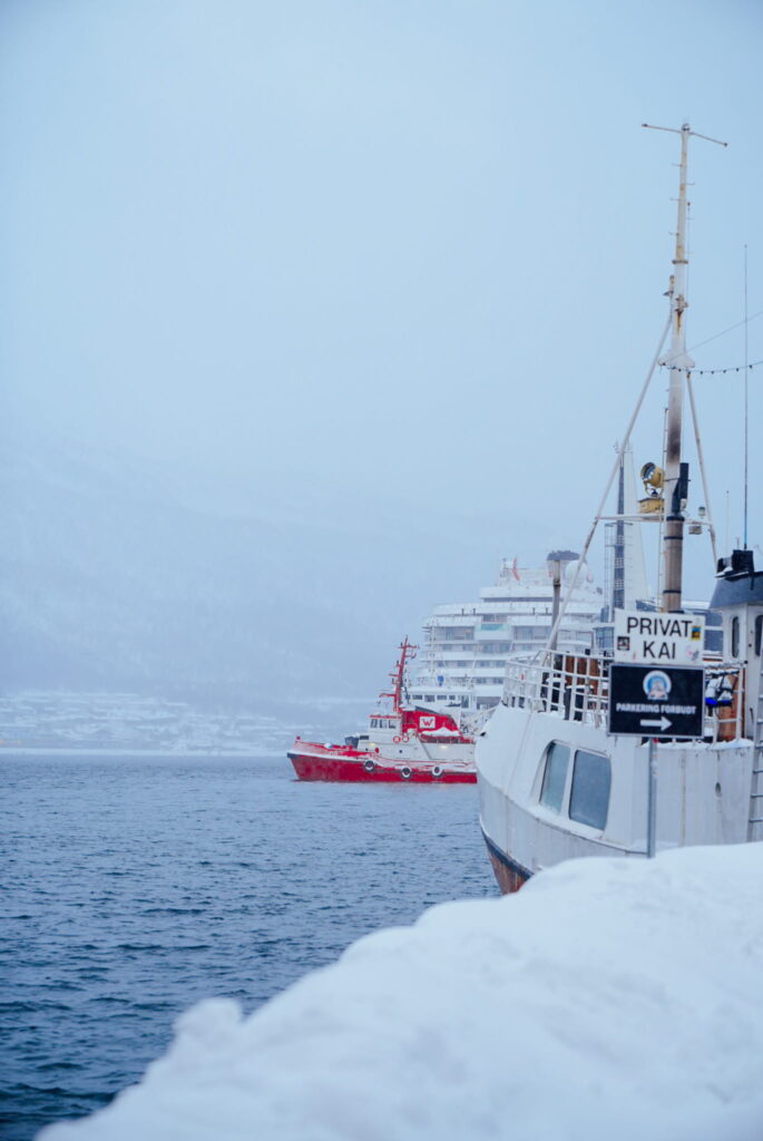 A snowy dock with a white boat in the foreground, a sign reading "PRIVATE KAI," and, as seen on a 3 Day Tromso Itinerary, a large red ship and white cruise ship in the misty, wintry background on calm water.