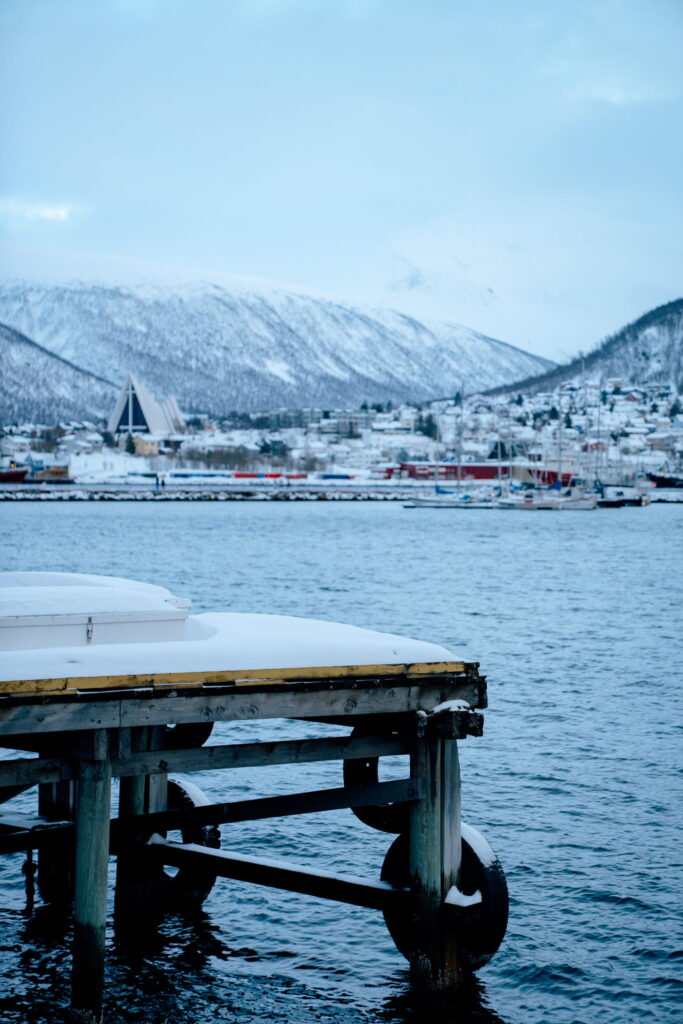 A wooden dock covered in snow extends over calm water, with snowy mountains, a modern triangular building, and a small town—perfect scenery for your 3 Day Tromso Itinerary—visible in the background under a cloudy winter sky.