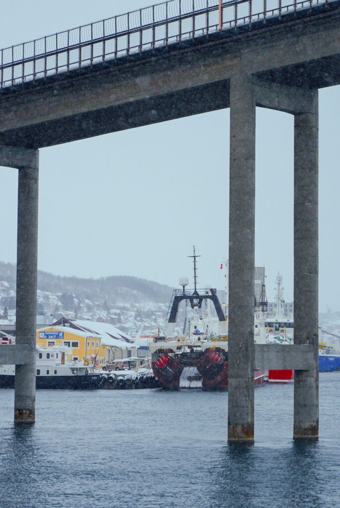 A large fishing boat docked at a snowy waterfront, framed between the concrete pillars of a bridge, sets the scene for your 3 Day Tromso Itinerary. Snow-covered hills and buildings rise in the background beneath a cloudy sky.