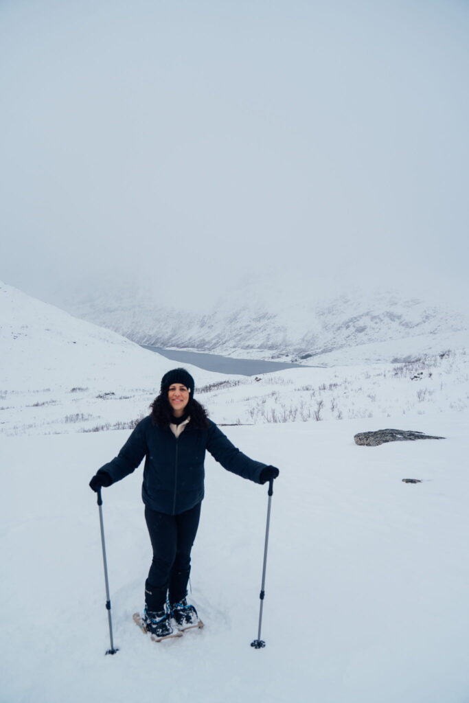 A person bundled in winter clothing stands on snowshoes, holding poles, in a snowy landscape with mountains and a cloudy sky—perfect inspiration for your 3 Day Tromso Itinerary.