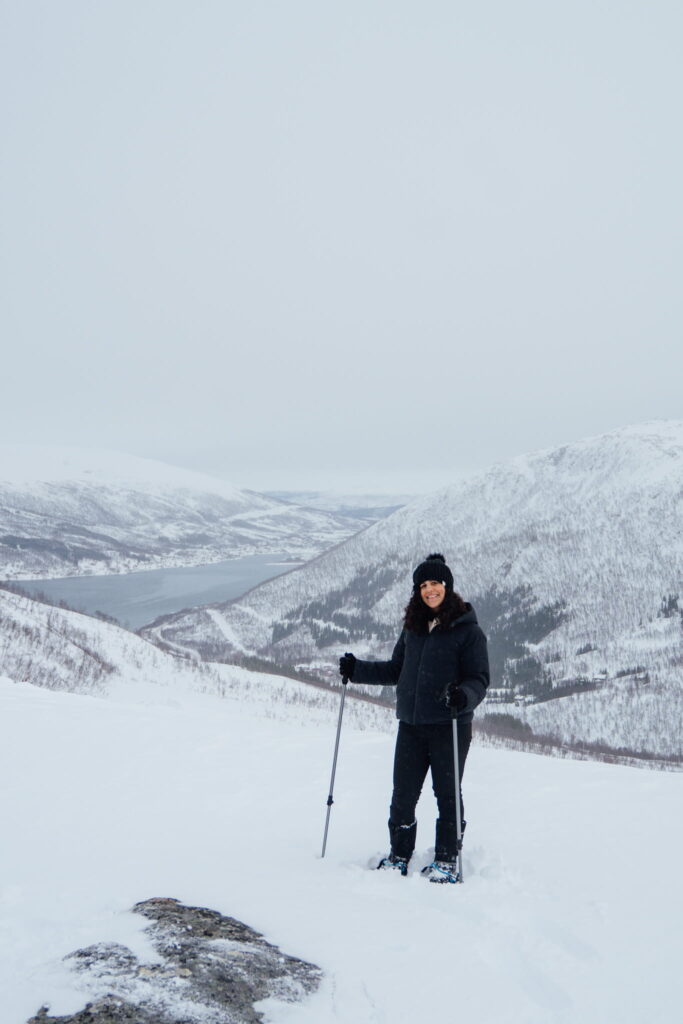 A person dressed in winter clothes and snowshoes stands on a snowy slope with trekking poles—an inspiring scene for any 3 Day Tromso Itinerary. Snow-covered mountains and a river valley stretch behind under an overcast sky.