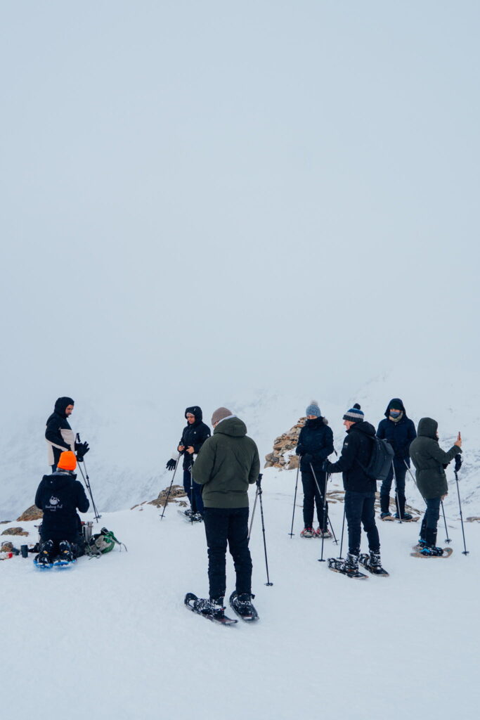A group of people dressed in winter clothing stand on a snowy mountain during their 3 Day Tromso Itinerary, wearing snowshoes and holding trekking poles. The sky is overcast, with a vast and foggy snowy landscape in the background.