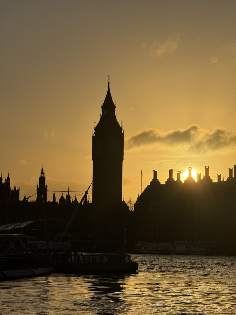 Silhouette of Big Ben and the Houses of Parliament in London at sunset, with the sky glowing orange and yellow behind the buildings—perfect for capturing on your sim local device, as reflections shimmer on the Thames River in the foreground.