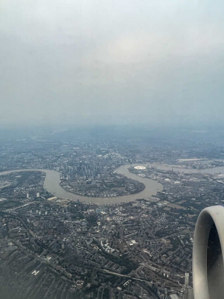 Aerial view of London with the River Thames curving through urban landmarks and a plane engine in the lower right corner. The overcast sky sets the scene—ideal for capturing your first sim local experience upon arrival.