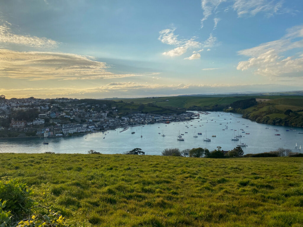 A scenic view of a coastal town with houses lining the waterfront, a river filled with anchored boats, green hills in the foreground, and a blue sky with scattered clouds at sunset—perfect for capturing memories to share with your sim local.